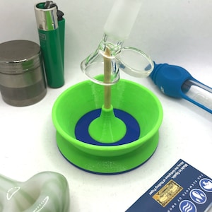May include: A green and blue 3D printed stand with a wooden dowel and a glass pipe. The stand is designed to hold a pipe upright. The text on the card reads "The Elements of Smoke, Alcoy, the birthplace of Rolling Paper, Made in Spain."