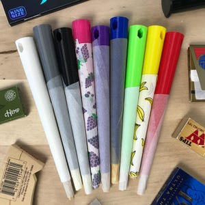 May include: A collection of pre-rolled cones in various colors, including white, gray, black, pink, purple, blue, green, yellow, and red. Some cones feature printed patterns like grapes and bananas. The image also shows boxes of tips and rolling papers.