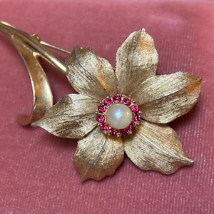 May include: A gold-toned flower brooch with textured petals and a central pearl surrounded by pink gemstones. The brooch has a curved stem and a pin closure, suitable for adding a touch of elegance to any outfit.