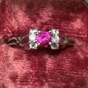 May include: A silver ring with a pink gemstone surrounded by smaller clear gemstones. The ring is set on a red velvet background.