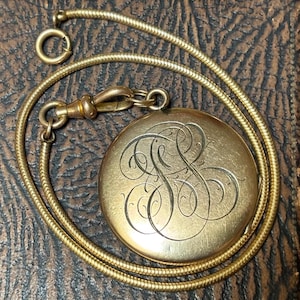 May include: A round gold-tone locket featuring an intricate, engraved monogram with swirling script letters on the front surface. The locket is attached to a flexible, gold-tone snake chain necklace that includes a circular spring ring clasp.