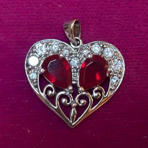 May include: A gold-toned heart pendant with two large, red gemstones and numerous small, clear crystals. The pendant has an ornate, filigree design and a loop for a chain. The background is a deep red color.