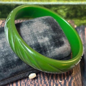 May include: A vibrant green bangle bracelet with carved leaf-like designs. The vintage jewelry piece is displayed on a dark, textured surface, showcasing its glossy finish and intricate details. The bracelet's color is a rich, saturated green.
