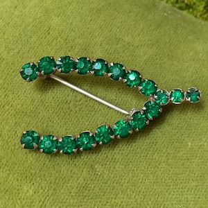 May include: A green rhinestone horseshoe brooch with a silver pin.