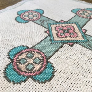 Puede incluir: Un dise&ntilde;o en forma de cruz bordado en tela blanca con hilo turquesa, rosa y beige. El dise&ntilde;o presenta cuatro motivos circulares con una forma de cruz central y un motivo en forma de diamante en el centro.
