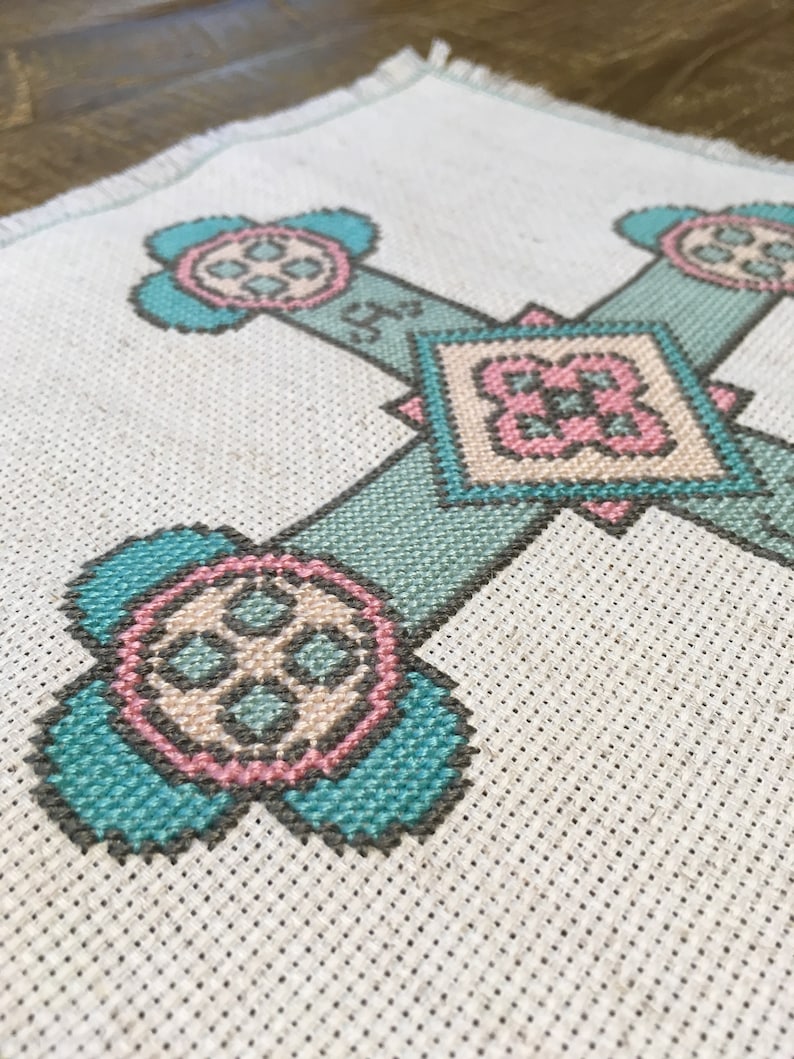 Puede incluir: Un dise&ntilde;o en forma de cruz bordado en tela blanca con hilo turquesa, rosa y beige. El dise&ntilde;o presenta cuatro motivos circulares con una forma de cruz central y un motivo en forma de diamante en el centro.