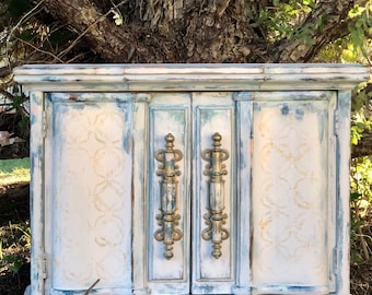 May include: A white painted wooden cabinet with two doors and gold-toned hardware. The cabinet has a distressed finish and a decorative design on the doors.