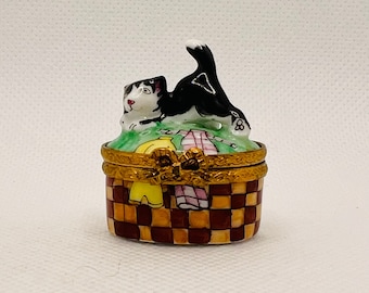 May include: A black and white cat figurine sits atop a small, oval-shaped porcelain box. The box is decorated with a checkered pattern in shades of brown and yellow. The cat is depicted in a playful pose, with its tail curled up and its head tilted to the side.