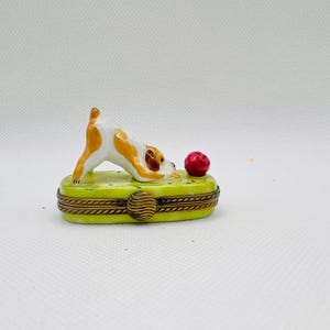 以下が含まれることがあります： 緑色の台座の上に、白と茶色の犬の置物が赤いボールを追いかけている、磁器製の小物入れです。