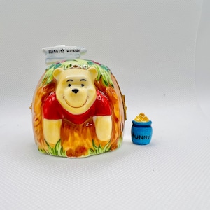 May include: A ceramic Winnie the Pooh figurine in the shape of a honey pot with a sign that reads "Rabbit's House". The figurine is painted in shades of brown, yellow, red, and green. A small blue ceramic honey pot with the word "Hunny" on it is next to the figurine.