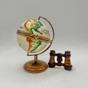 May include: A decorative globe with a hinged lid, featuring a map of the world with continents in green, orange, and yellow. The globe is mounted on a brass stand with a wooden base. A miniature pair of brown binoculars is next to the globe.