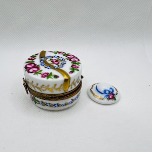 May include: A small, round porcelain trinket box with a floral design and gold accents. The lid features a blue ribbon and a floral design. The box is closed.