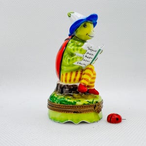 Op de afbeelding: Een porseleinen sieraden doosje met een lieveheersbeestje dat een blauw-witte hoed draagt, op een groene stronk zit en een boek leest. De doos is versierd met groene, gele en rode details. Een klein lieveheersbeestje staat op de tafel naast de doos.