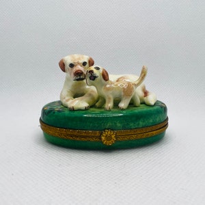 以下が含まれることがあります： 白いラブラドールレトリバーの犬2匹が描かれた磁器製の小物入れ。大きな犬は横たわっており、頭を小さい犬の方に向けている。小さい犬は後ろ足で立っている。箱は緑色で、金色の縁取りと花柄の模様が施されている。
