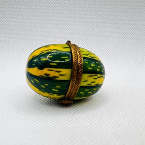 Könnte beinhalten: Eine kleine, ovale Schmuckschatulle mit einem grün-gelben Streifenmuster. Die Schachtel ist aus Porzellan und hat einen goldfarbenen Metallrand.