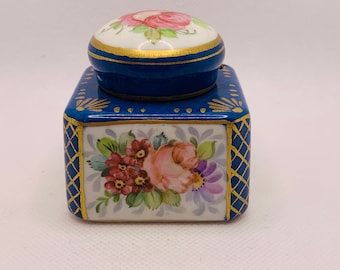 Puede incluir: Un tintero cuadrado de porcelana con un diseño floral y ribetes dorados. La tapa es redonda y tiene un diseño floral con acentos rosados y verdes.
