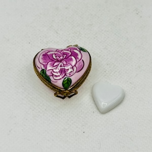 Puede incluir: Una pequeña caja de corazón con un diseño floral rosa y verde. La caja está hecha de porcelana y tiene una bisagra de metal dorado.