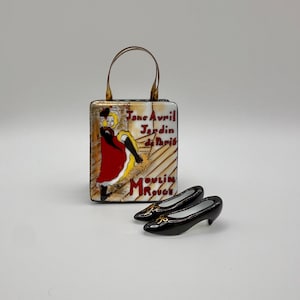 Puede incluir: Un bolso decorativo con asa, con un diseño de cartel vintage del Moulin Rouge. El bolso se combina con un par de zapatos de tacón negros. El arte del cartel incluye a una mujer con un vestido rojo y el texto "Jane Avril Jardin de Paris Moulin Rouge."