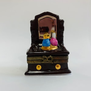 May include: A small, dark brown ceramic vanity with a mirror, two drawers, and gold accents. The vanity has a blue and pink bottle on top.