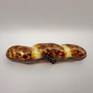 May include: A peanut-shaped decorative box featuring a glossy, mottled brown and cream tortoiseshell-like finish. The object has a metallic, ribbed base and a small, dark, textured decorative element attached to the side of the central section.