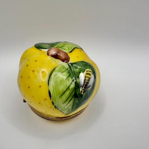 Puede incluir: Una caja de baratijas de cerámica amarilla con forma de manzana, con un diseño de hoja verde y abeja. La caja tiene una base de metal dorado y un tallo marrón. La manzana tiene pequeñas manchas marrones.