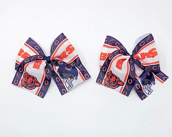 May include: Two white hair bows with a navy blue and orange Chicago Bears logo print. The bows feature the team's logo, the words "Bears" and a football pattern.