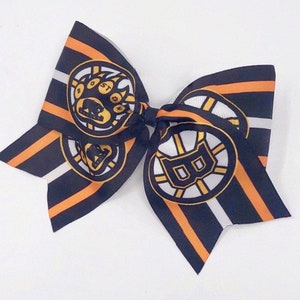May include: A black and orange striped hair bow with a white circle and the Boston Bruins logo. The logo features a bear paw print and the letter "B".