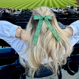 May include: A woman with blonde hair wearing a white shirt and a green and gold bow in her hair. The bow has the words "Packers" repeated on it.