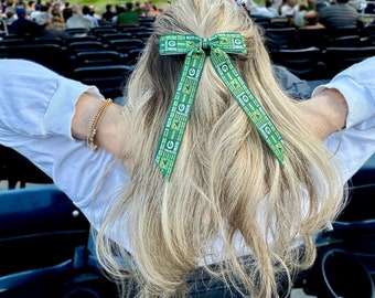 Puede incluir: Una mujer con cabello rubio que lleva una camisa blanca y un lazo verde y dorado en el cabello. El lazo tiene las palabras "Packers" repetidas.
