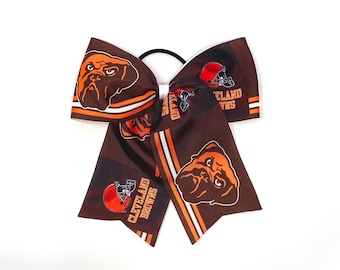 Pode incluir: Laço de cabelo marrom e laranja com o logotipo do Cleveland Browns e um bulldog como mascote. O laço tem listras brancas e o texto "Cleveland Browns".