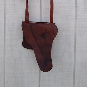 May include: A brown leather holster with a long strap, designed to be worn over the shoulder. The holster is a deep, rich brown color and appears to be handmade. The strap is thin and adjustable, and the holster has a unique shape.