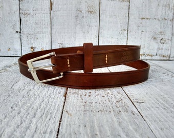 May include: A brown leather belt with a silver buckle and a decorative design on the strap. The belt is rolled up and lying on a white wooden surface.