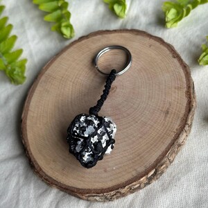 May include: A black and white speckled stone keychain with a black woven cord and a silver-colored keyring. The stone is wrapped in a macrame-style weave. The keychain rests on a wooden slice.