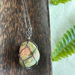 May include: A handcrafted necklace featuring a large, oval unakite stone pendant. The stone displays a mix of green and pink hues, encased in a macrame net and suspended from a braided cord. The necklace rests on a dark wooden surface.