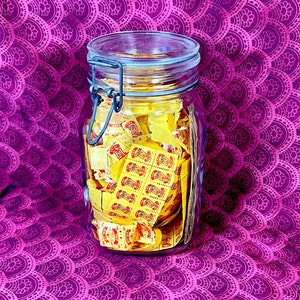 Puede incluir: Un tarro de vidrio lleno de boletos de papel amarillos. El tarro tiene una tapa de metal y un cierre de metal. El tarro está sentado sobre una tela estampada de color morado y blanco.