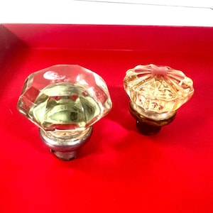Peut inclure: Deux boutons de porte anciens en verre. L'un est transparent avec un design à facettes et une base argentée. L'autre est ambré avec un motif en étoile et une base marron foncé. Les deux sont posés sur un fond rouge vif.