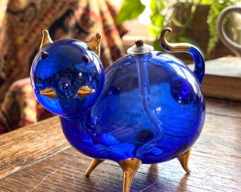 Könnte beinhalten: Eine kobaltblaue Öllampe aus Glas in Form einer Katze. Die Lampe hat goldfarbene Glasakzente an den Ohren, der Nase, dem Schwanz und den vier Füßen. Ein kleiner Dochtträger aus Metall befindet sich oben auf dem abgerundeten Körper.