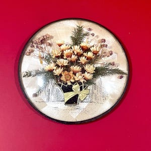 Puede incluir: Arte mural redondo con un arreglo floral seco. El arreglo incluye flores de color crema, ramitas verdes y acentos secos, todo en un jarrón negro y dorado con un lazo. El arte está encerrado en un marco circular.