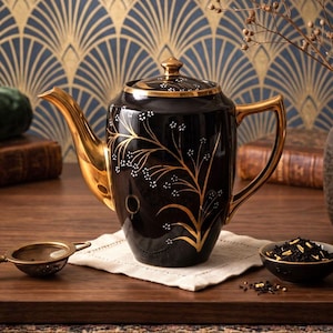 Könnte beinhalten: Eine schwarze Keramik-Teekanne mit goldfarbenem Griff, Ausguss und Deckelrand. Der Körper ist mit einem goldenen botanischen Zweigmuster verziert, das durch kleine weiße Punkte akzentuiert wird. Die Teekanne hat einen passenden Deckel mit goldenem Knauf.
