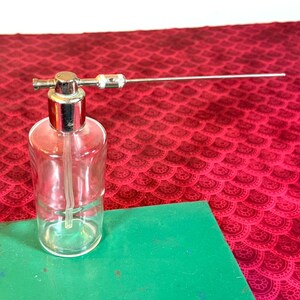 May include: A clear glass bottle with a silver metal top and a long, thin metal tube extending from the top. The bottle sits on a green surface against a red patterned background. The bottle has a clear plastic tube inside.