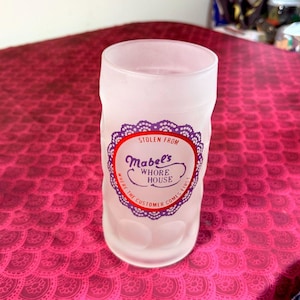 May include: Frosted glass with a decorative label. The label reads "Stolen From Mabel's Whore House" in a circular design with a purple lace border and red accents. The text "Where the customer comes first" is also included.