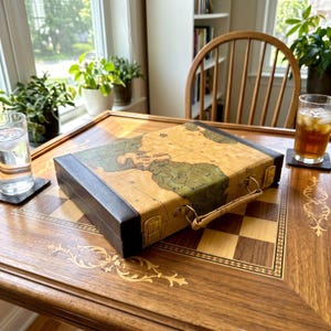 May include: A rectangular carrying case featuring a vintage-style map print on the top surface, accented with dark brown leather-like trim, metal latches, and a matching handle, resting on a wooden table with a checkered inlay pattern.