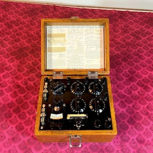 Puede incluir: Un antiguo juego de pruebas Leeds & Northrup en una caja de madera abierta. El interior presenta un panel de control negro con múltiples diales, interruptores y un medidor. La tapa muestra instrucciones y diagramas.