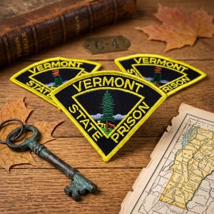 May include: Three triangular embroidered patches featuring a black background with a yellow border and text reading VERMONT STATE PRISON. The center of each patch displays a green pine tree above a small brown animal on a light blue base.