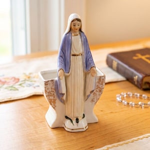 Könnte beinhalten: Eine Keramikfigur der Jungfrau Maria, die vor einem kleinen, oben offenen Gefäß steht. Die Figur trägt ein weißes Gewand und einen hellblauen Umhang mit Goldrand. Das Gefäß hat eine weiße Basis mit goldenen Sprenkeln an den Seiten.