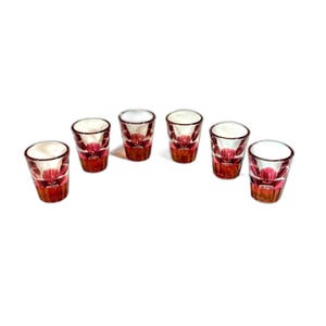 May include: A set of six small drinking glasses featuring a clear upper section and a vibrant pink base with a faceted, starburst-like pattern at the bottom. Each glass has a cylindrical shape with a thick, weighted base and smooth rim.