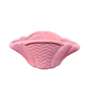 Könnte beinhalten: Ein rosa Keramikkorb mit gewelltem Rand und Zickzackmuster auf der Vorderseite. Der Korb hat eine geschwungene Form und ist zur Aufnahme von Gegenständen bestimmt. Die Farbe ist ein weiches, pastellfarbenes Rosa.