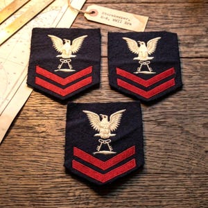 Puede incluir: Tres parches bordados azul marino con un águila blanca sobre dos galones rojos. Una etiqueta de papel beige muestra el texto Storekeeper, E-6, WWII Era. Los parches tienen forma de escudo con base puntiaguda y costuras blancas.