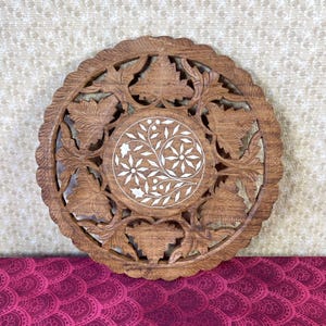 May include: A round wooden wall decor with intricate carvings of leaves and flowers. The center of the decor features a circular inlay of white mother of pearl with a floral design.