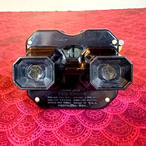 Puede incluir: Un visor de estereoscopio Vision Master negro vintage. El visor tiene dos oculares y un área de visualización central. El texto en la parte inferior dice "Vision Master" y "Portland, Ore."